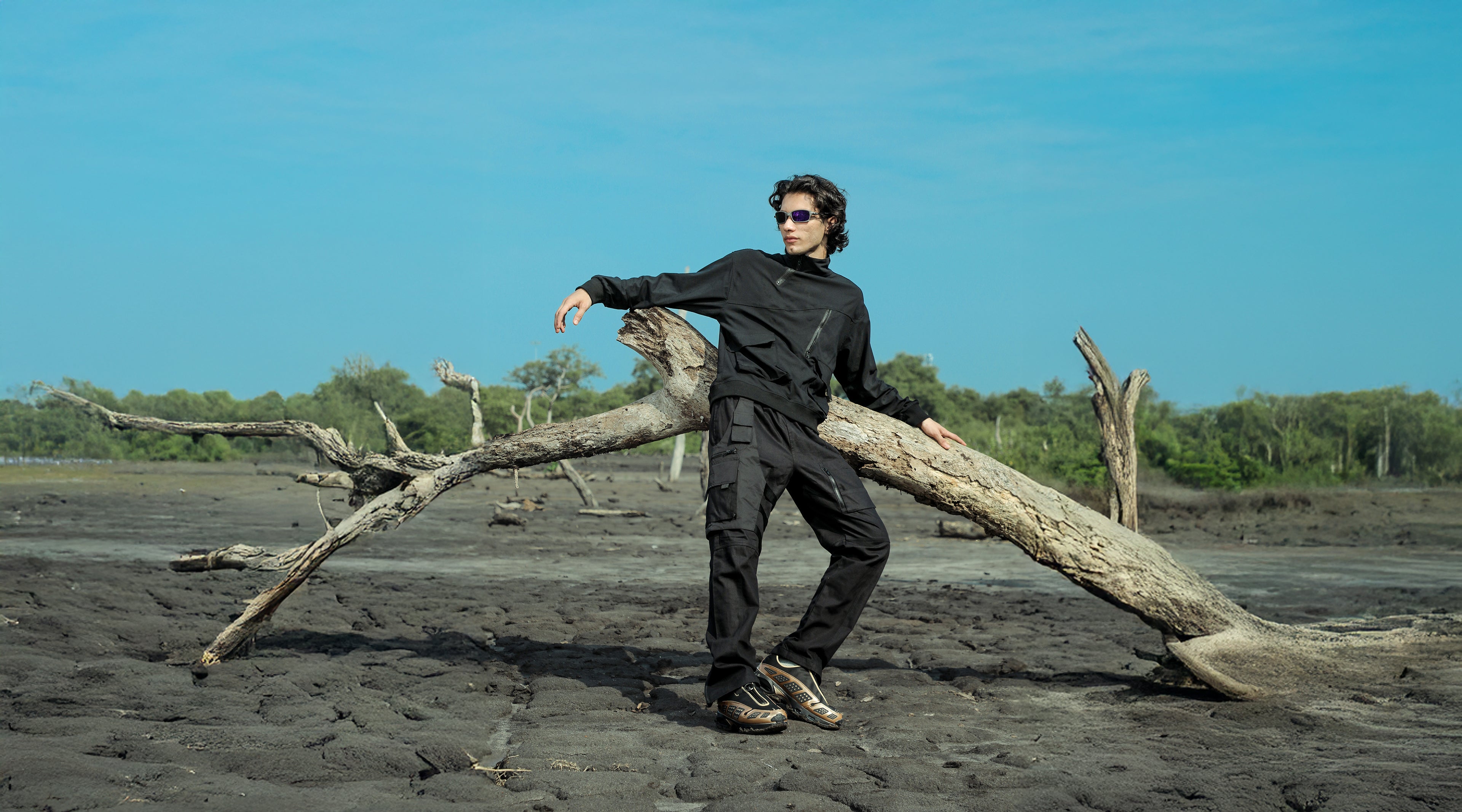 Person in black outfit standing on a beach with a large log, wearing sunglasses.